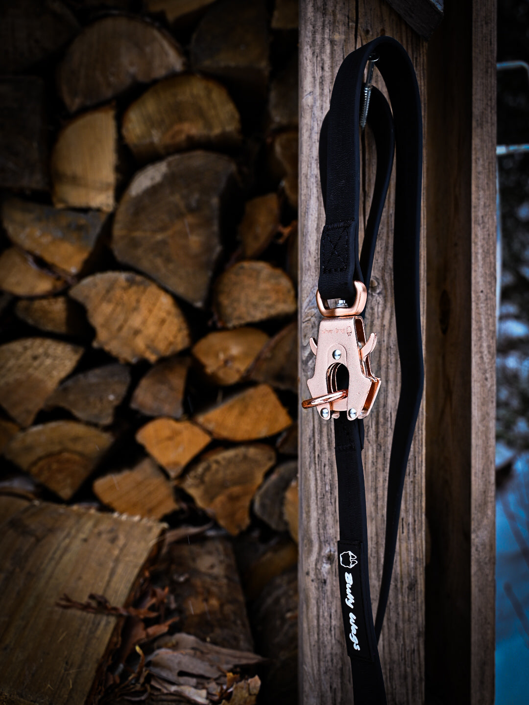 Black PVC dog leash with rose gold hardware, hanging from a wooden post with a stacked woodpile in the background.